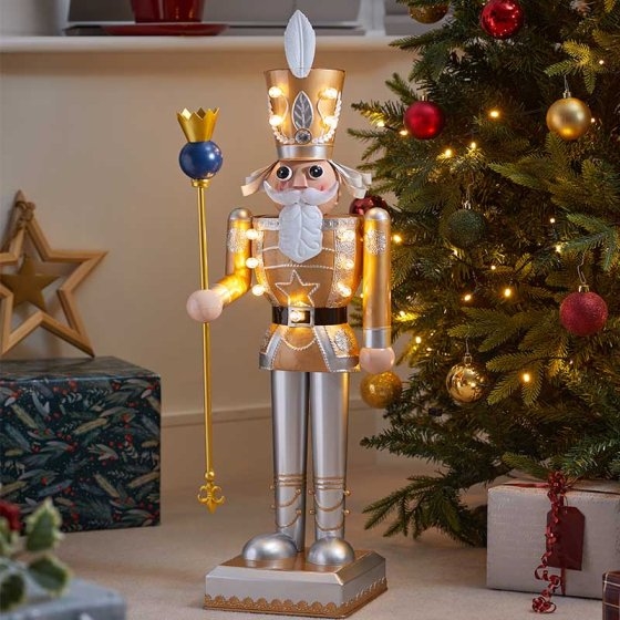 Gold and white nutcracker with a crown and scepter, featuring LED lights, standing beside a decorated Christmas tree and gifts.