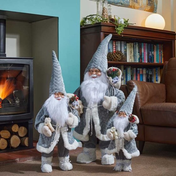 Three grey, plush Santa figures with white beards and hats, adorned with floral accents, stand by a cozy fireplace in a warmly decorated room.