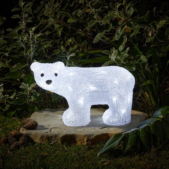 A glowing, translucent white bear sculpture with embedded LED lights, set on a stone surface surrounded by greenery and pinecones.