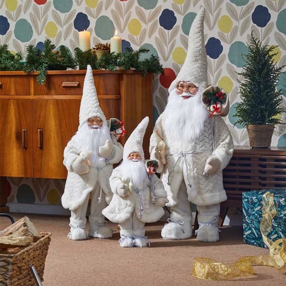 Three cream-colored Papa Noel figures with long white beards and pointed hats, adorned with holly, stand beside a wooden cabinet and festive decorations.