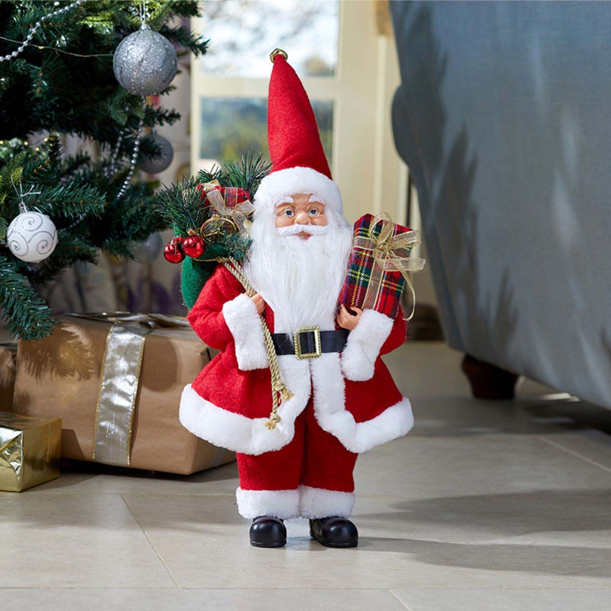 Festive Santa figure in a red suit with white trim, holding a sack and gifts, featuring a long white beard and a tall red hat, set near a Christmas tree and wrapped presents.