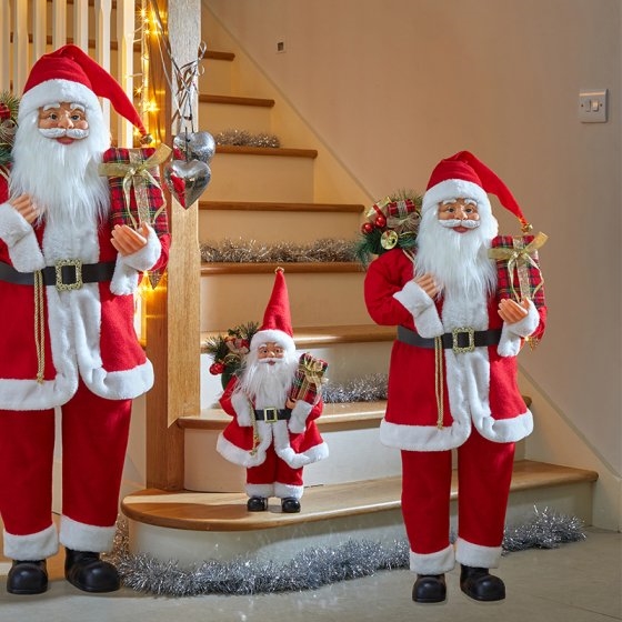 Three plush Santa figures in red and white, with long white beards, holding gifts, positioned on a staircase decorated with tinsel and lights.