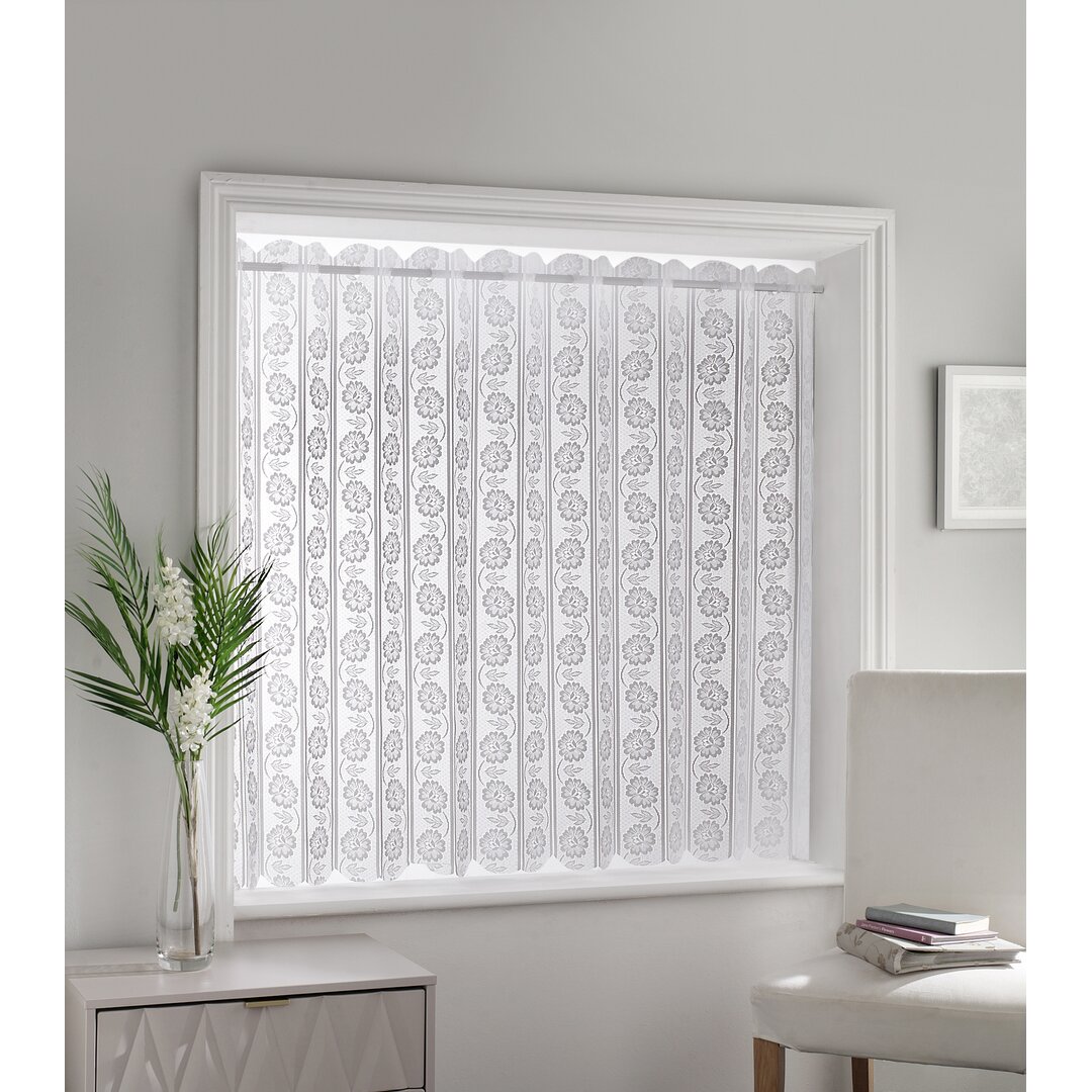 White floral lace louvre style blind featuring intricate patterns, hanging vertically in a window frame, complemented by a modern chair and a decorative plant nearby.