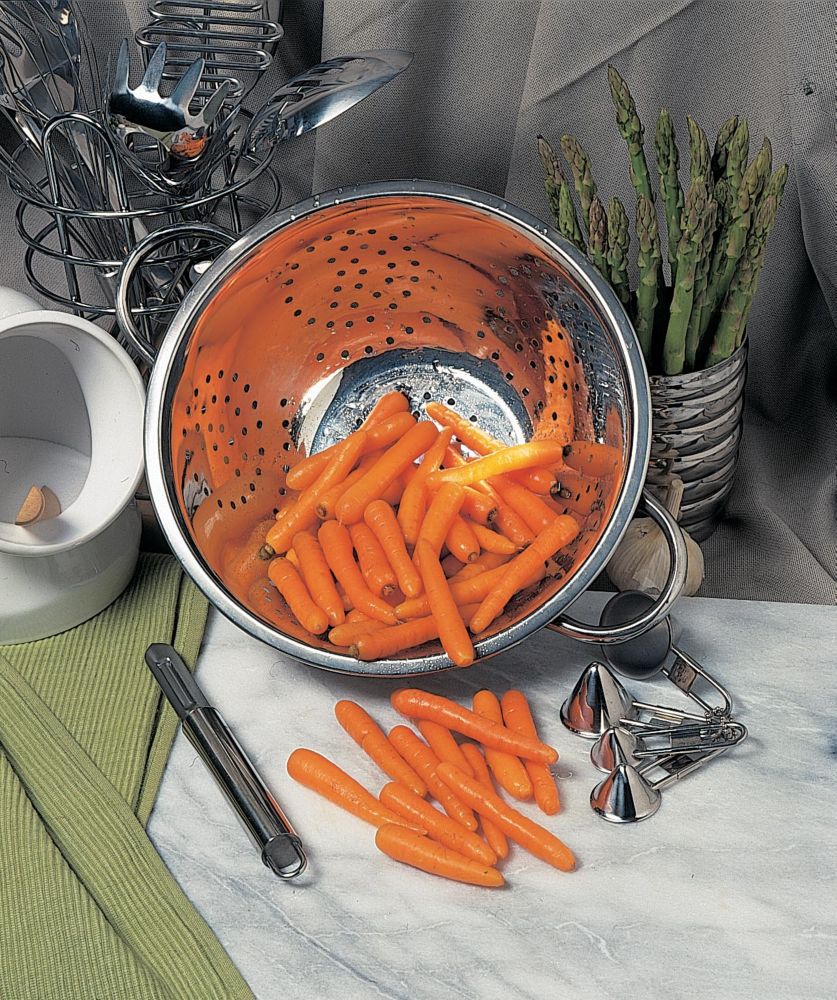 A colander filled with vibrant orange carrots sits on a marble surface, surrounded by kitchen utensils and fresh asparagus, with a green cloth underneath.