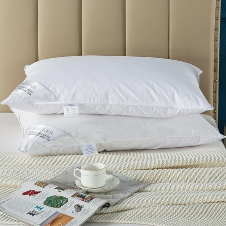 Two plush white pillows made from natural Hungarian goose feathers and down, resting on a bed with a textured cream blanket and a magazine beside a cup on a saucer.