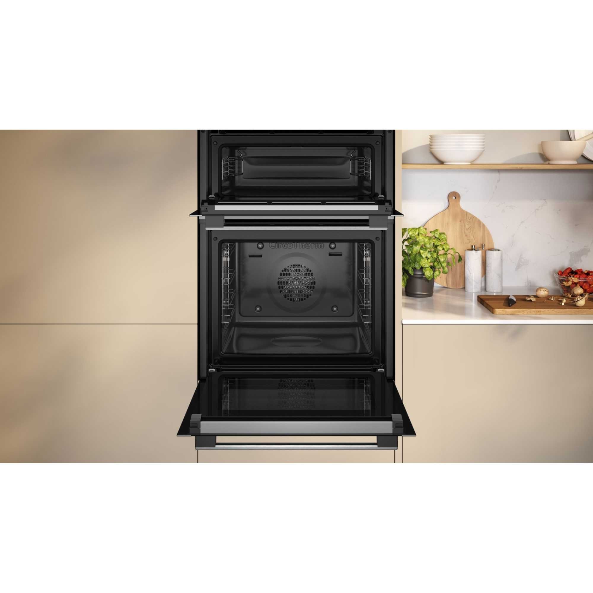 Built-in double oven in graphite finish, featuring a sleek design with a self-cleaning function, glass door, and modern control panel, set against a light kitchen backdrop.