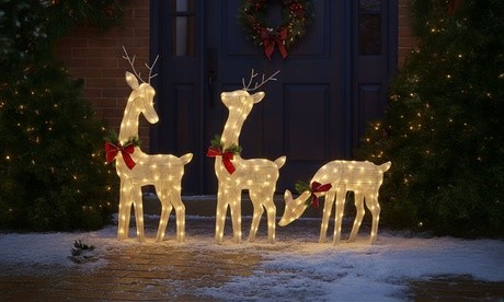 Three illuminated reindeer decorations in white, adorned with red bows and green garlands, standing on a snowy pathway near a festive door with a wreath.