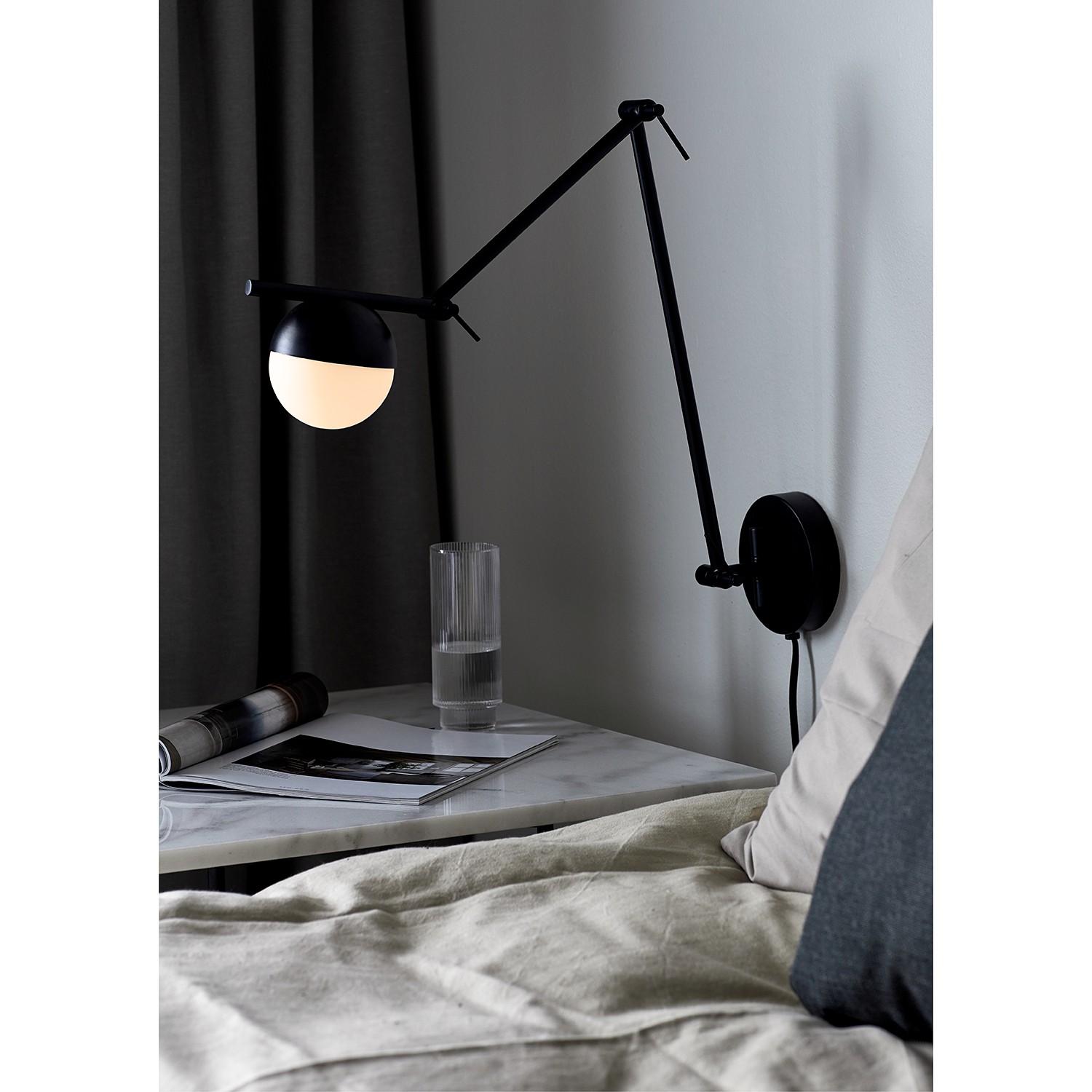 Nordlux Contina Wall Light in black features a sleek, adjustable arm and a round, white glass shade, mounted on a circular wall plate, beside a bedside table with a glass and magazines.