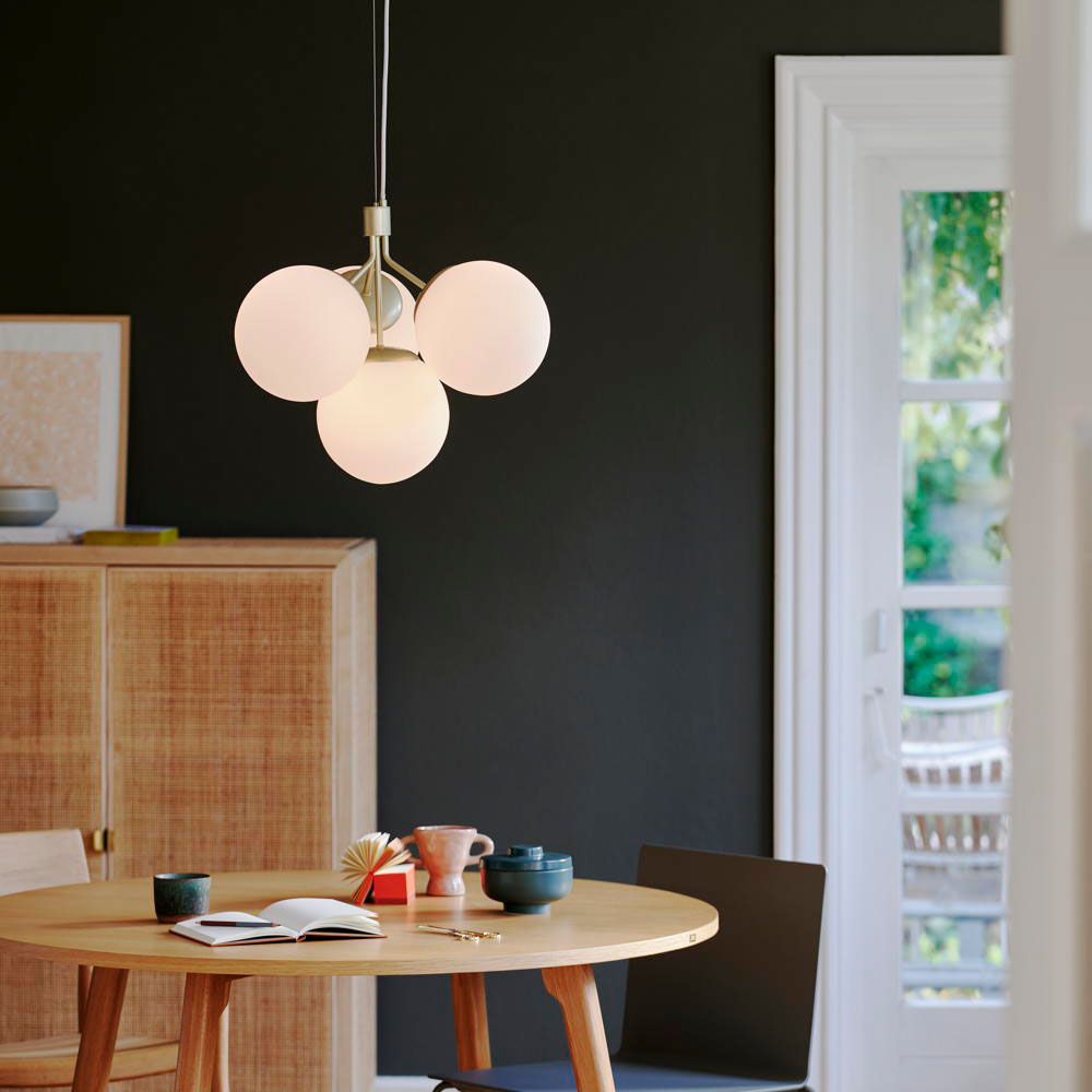 Brass pendant light with four spherical white glass shades, hanging from a sleek cord, illuminating a modern dining space with a wooden table and dark walls.