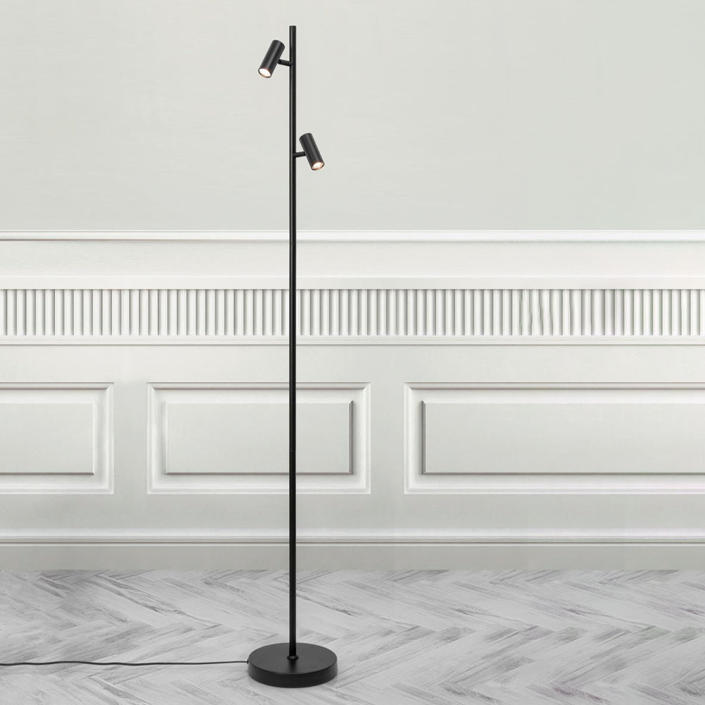Sleek black floor lamp with two adjustable cylindrical light fixtures on a slender pole, set against a light-colored wall and herringbone-patterned floor.