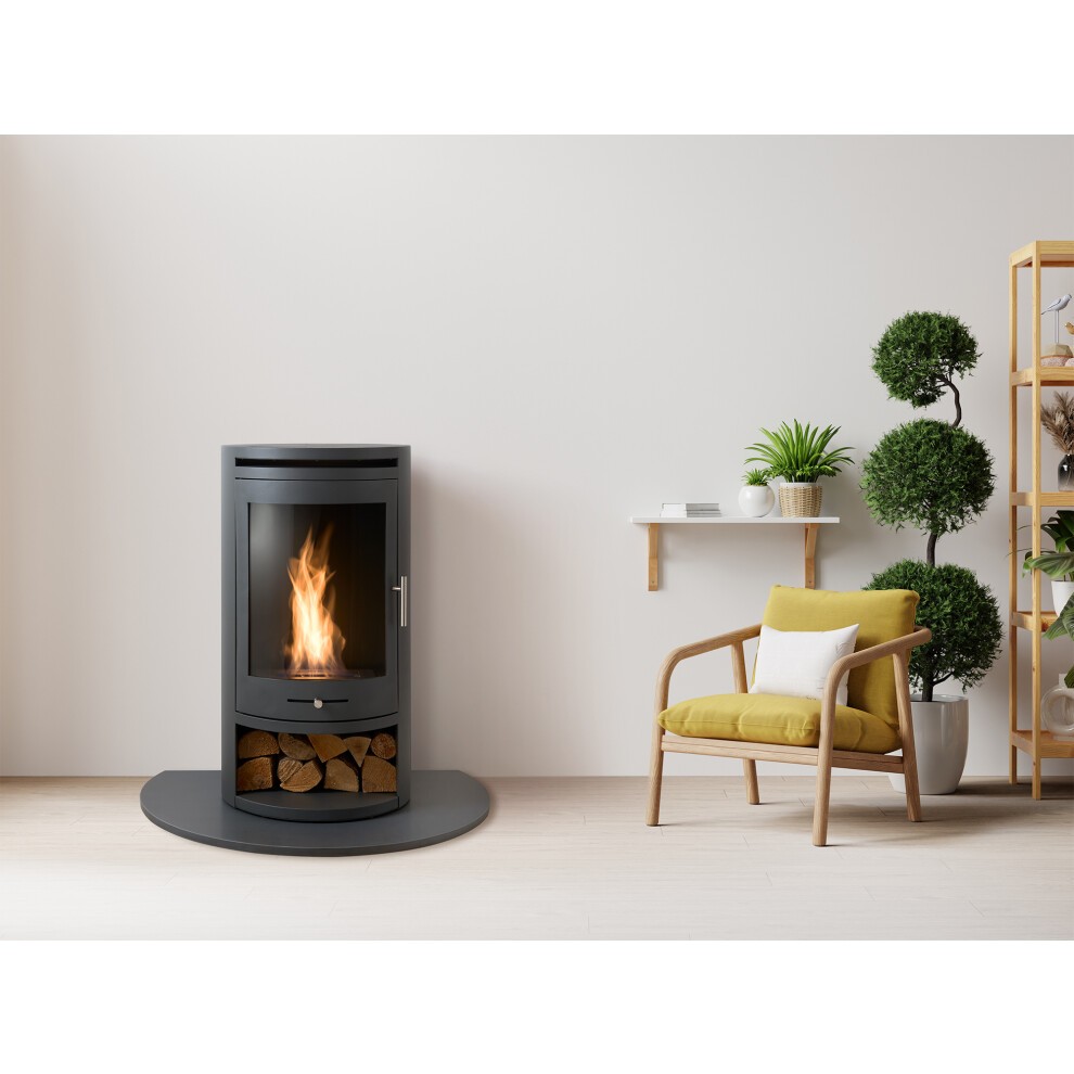 Charcoal grey OKO S5 bioethanol cylinder stove with a glass front, log storage compartment, and flames visible, positioned next to a modern chair and greenery.
