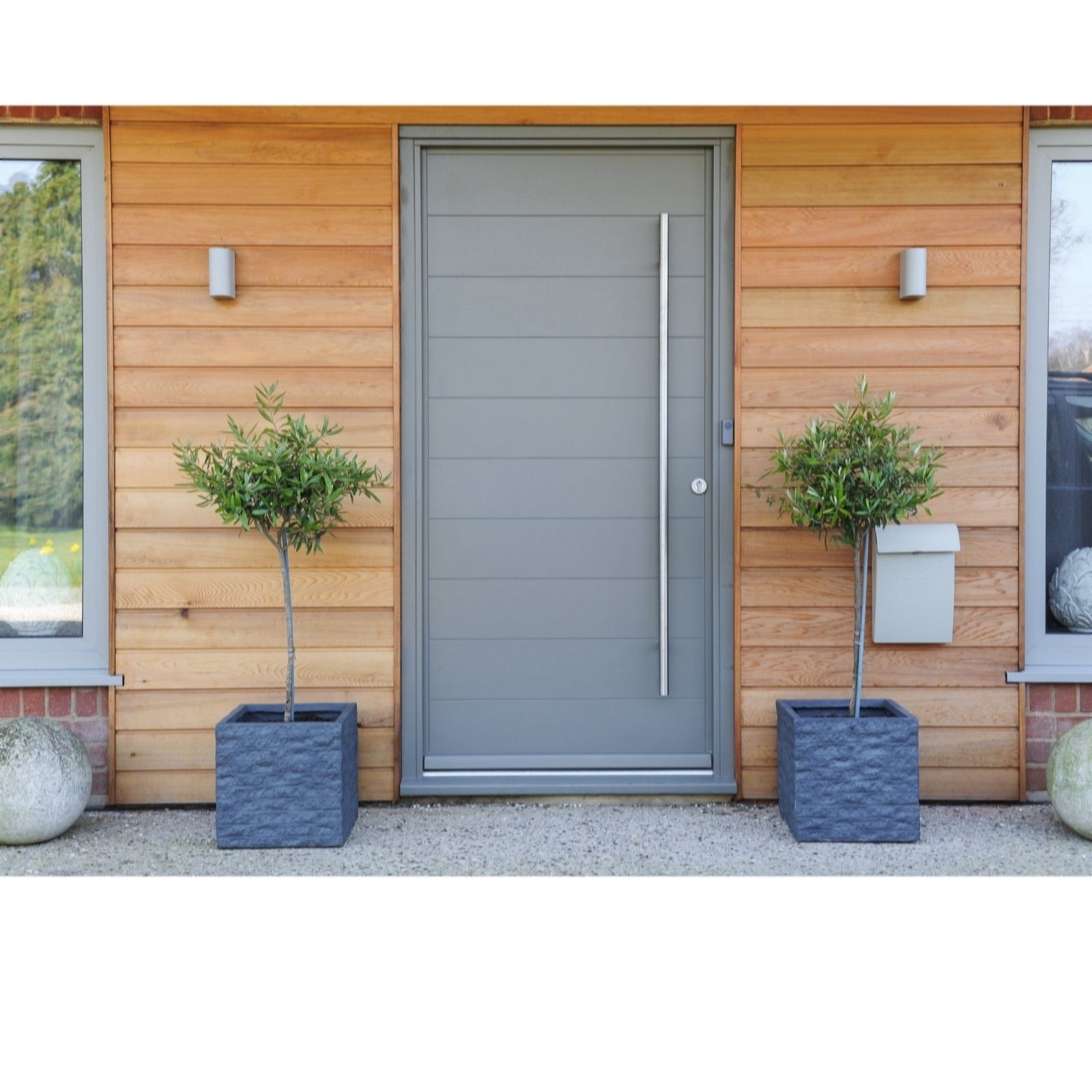 Two potted Olea Europaea plants in textured grey planters flanking a modern grey front door with a sleek vertical handle, set against a wooden wall.