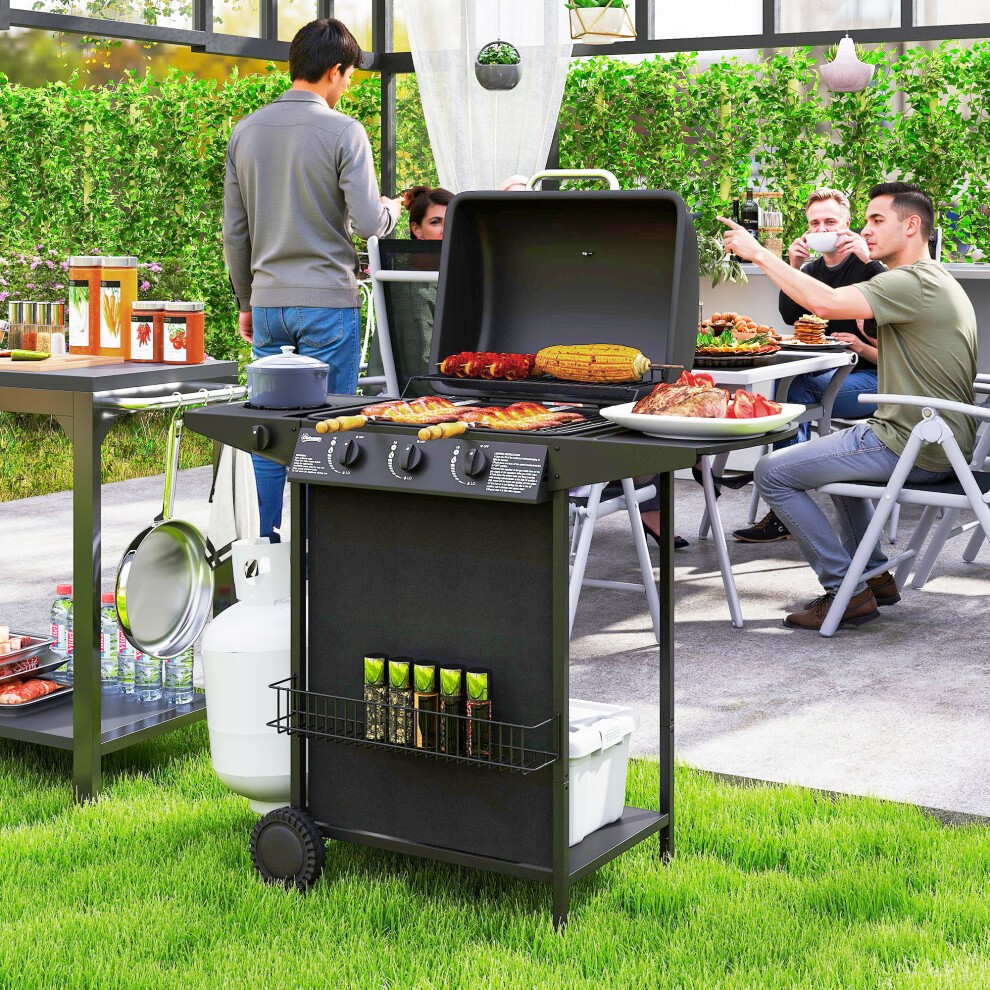 Portable black gas barbecue grill with four burners, a lid, and side shelves, set on wheels for easy mobility, surrounded by people enjoying a garden gathering.