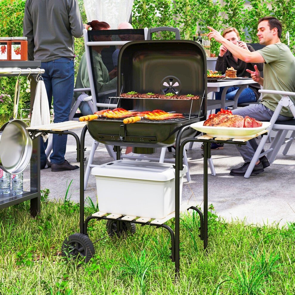 Portable black charcoal barbecue grill with a lockable lid, side shelves, and a vent, set on wheels, surrounded by people enjoying a meal outdoors.