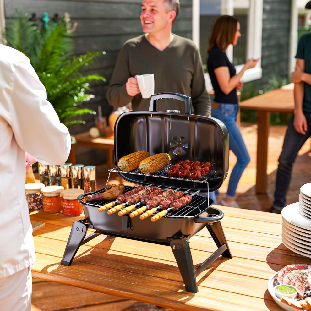 Portable black charcoal barbecue grill with a folding design, featuring a cooking grate with skewers and corn, set on a wooden table with people socializing in the background.