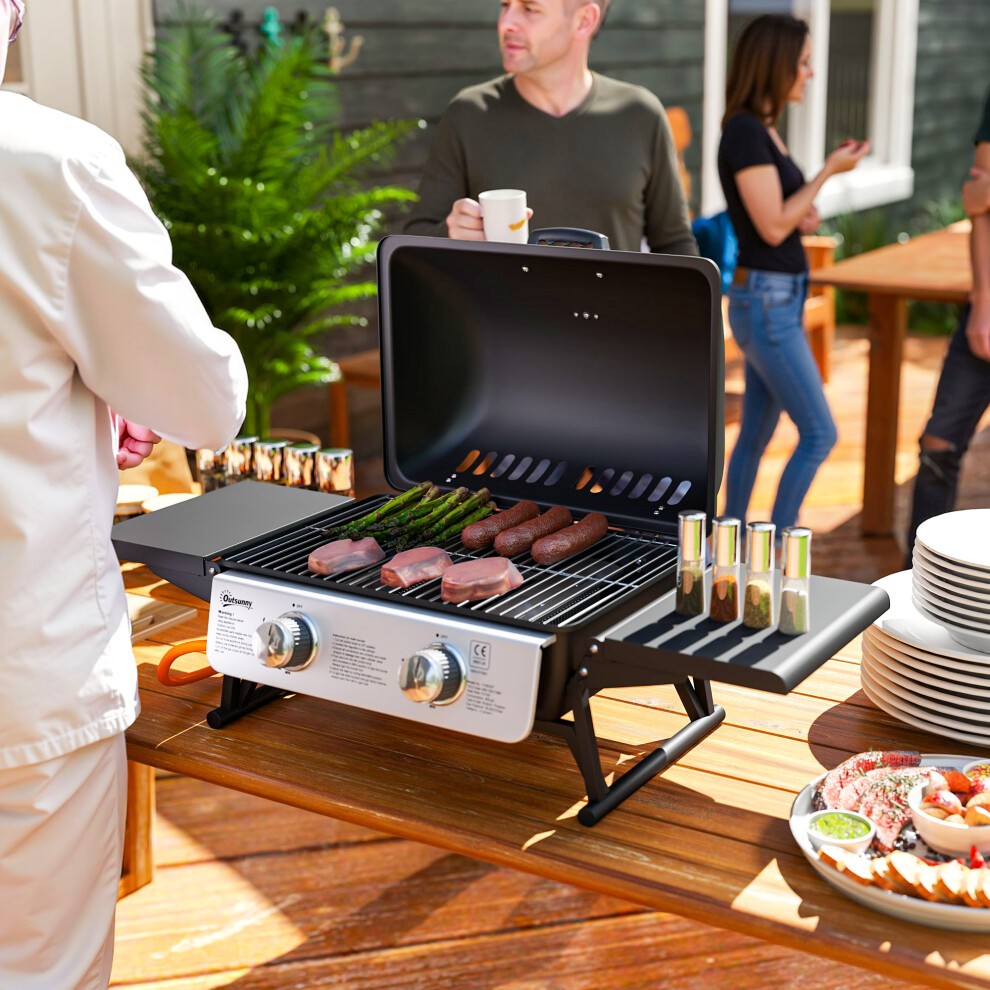 Portable black gas barbecue grill with a thermometer, featuring a double burner, metal grates, and a spacious cooking area, set on a wooden table with food and people in the background.