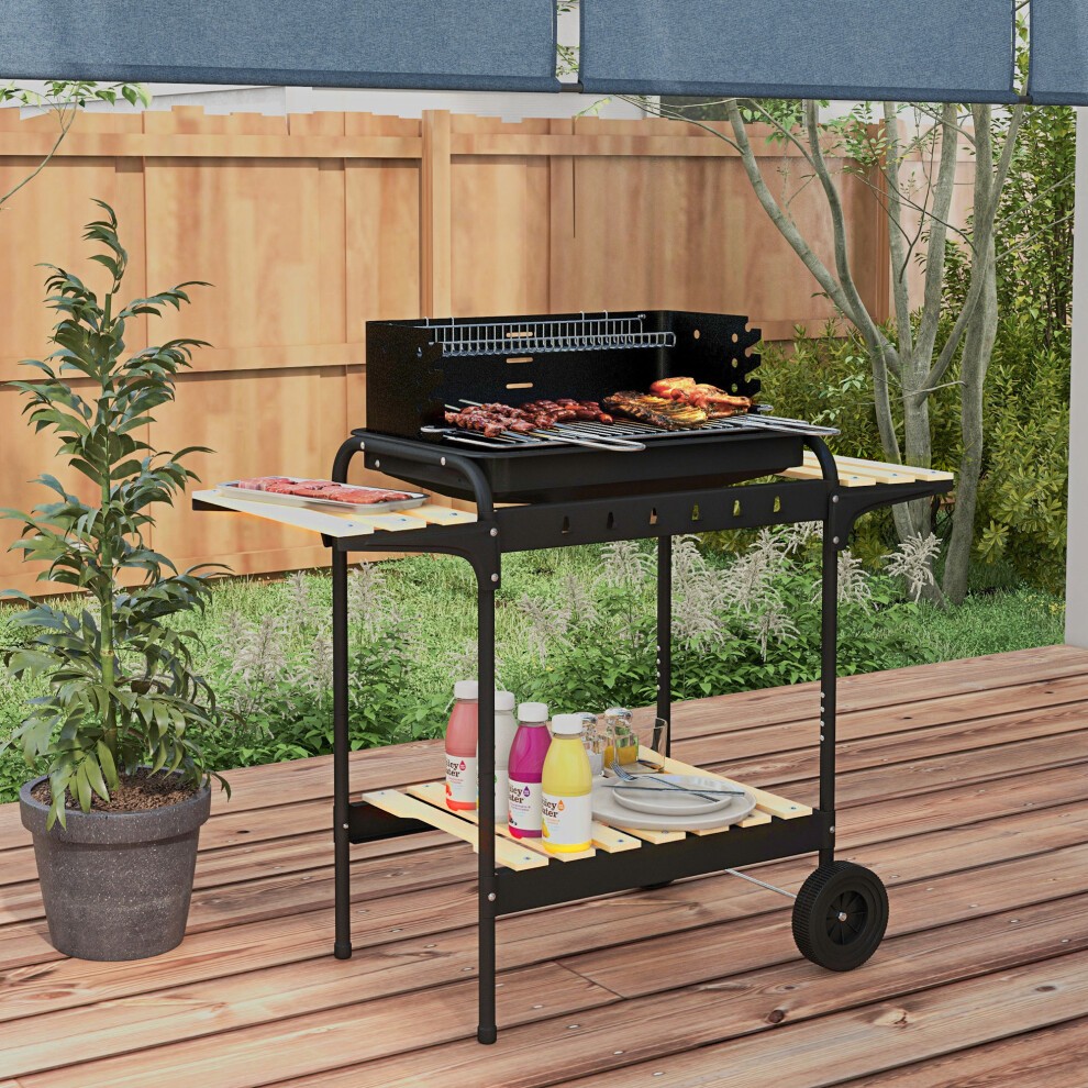 Black charcoal barbecue grill trolley with five adjustable grill heights, ash catcher, and wooden side shelves, set in a garden with greenery and a wooden fence.