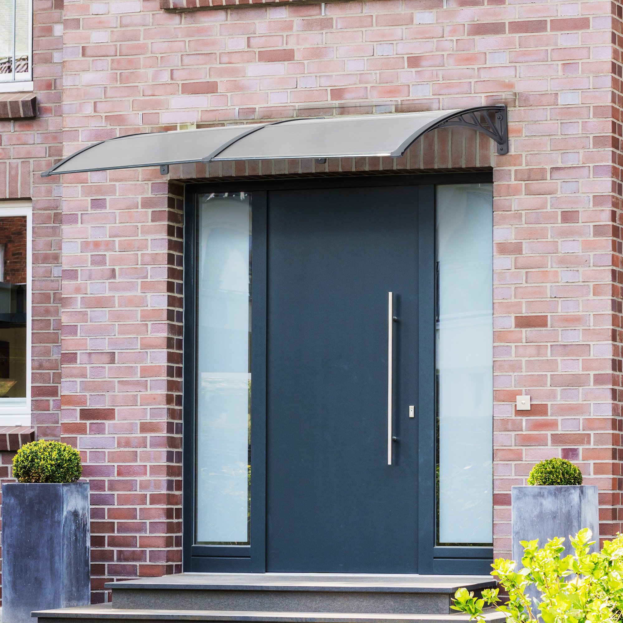 Clear polycarbonate awning with a curved design, mounted above a dark front door, providing shelter from rain, complemented by brick walls and potted plants on either side.