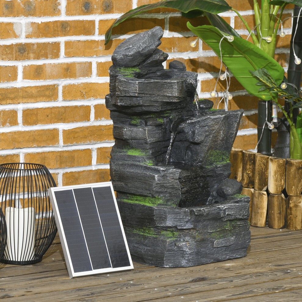 Dark grey stone-like garden fountain with cascading water feature, LED light, surrounded by greenery, and a solar panel on a wooden deck.