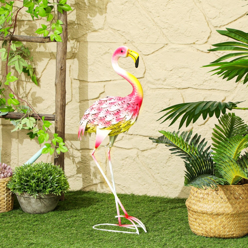 Colorful pink and yellow metal flamingo garden statue with intricate feather detailing, standing on a white stake amidst green plants and textured beige wall.