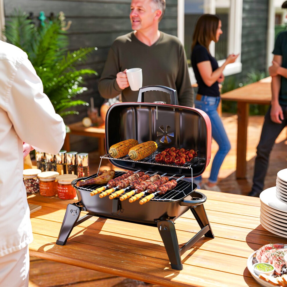 Red Outsunny folding charcoal barbecue grill with a black base, featuring a spacious cooking area with skewers and corn, set on a wooden table outdoors.