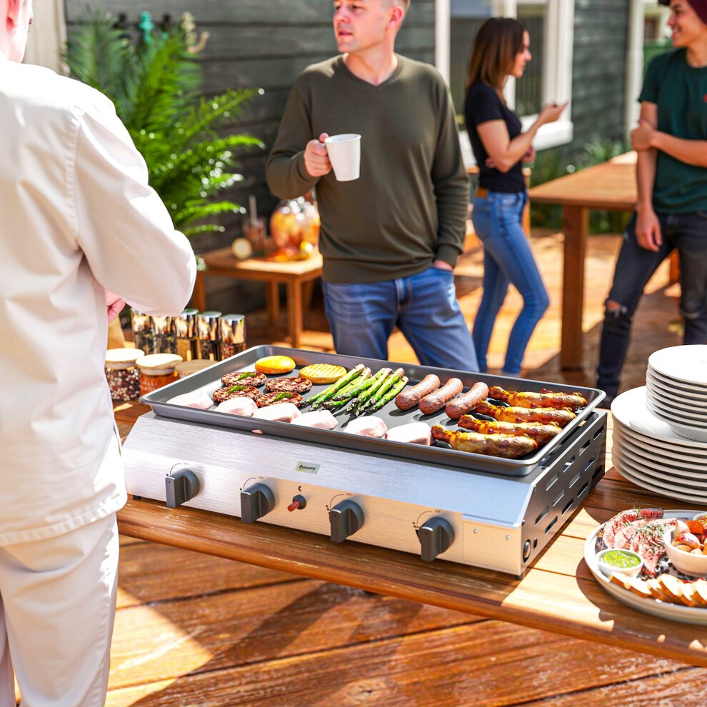 Silver-tone gas grill with a non-stick griddle, featuring multiple burners, cooking various meats and vegetables outdoors, surrounded by people enjoying a gathering.
