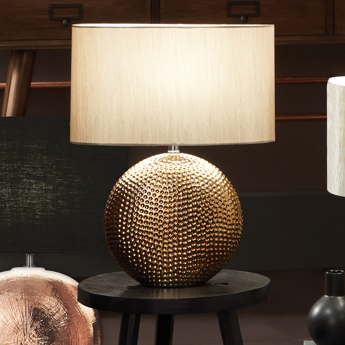 Ruma Dot Pattern Embossed Silver Textured Ceramic Table Lamp features a bronze spherical base with a dimpled design and a cream fabric shade, set on a dark wooden table.
