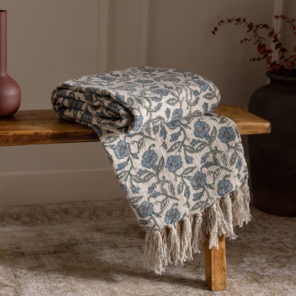 100% cotton floral printed throw in natural tones, featuring blue and green flowers, fringed edges, draped elegantly over a wooden bench.