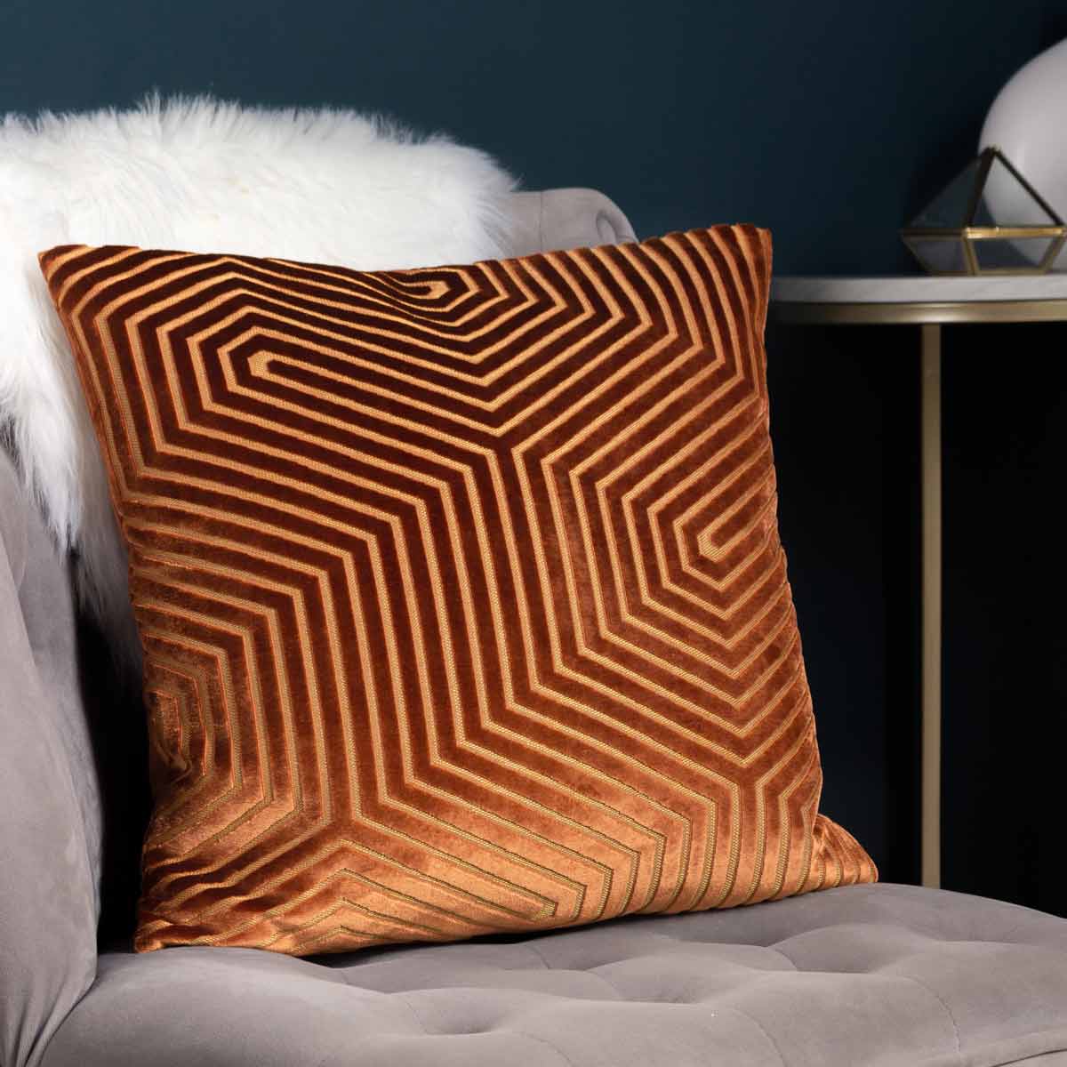 Red geometric cut velvet cushion featuring a striking maze-like pattern, measuring 45cm x 45cm, displayed on a soft gray chair with a faux fur throw.