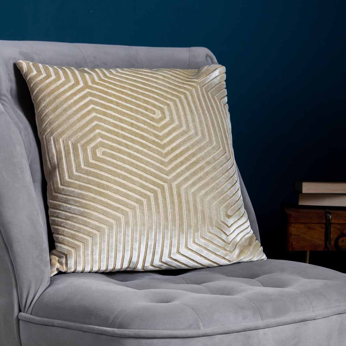 Ivory geometric cut velvet cushion with a textured zigzag pattern, measuring 45cm x 45cm, placed on a grey upholstered chair against a dark blue wall.