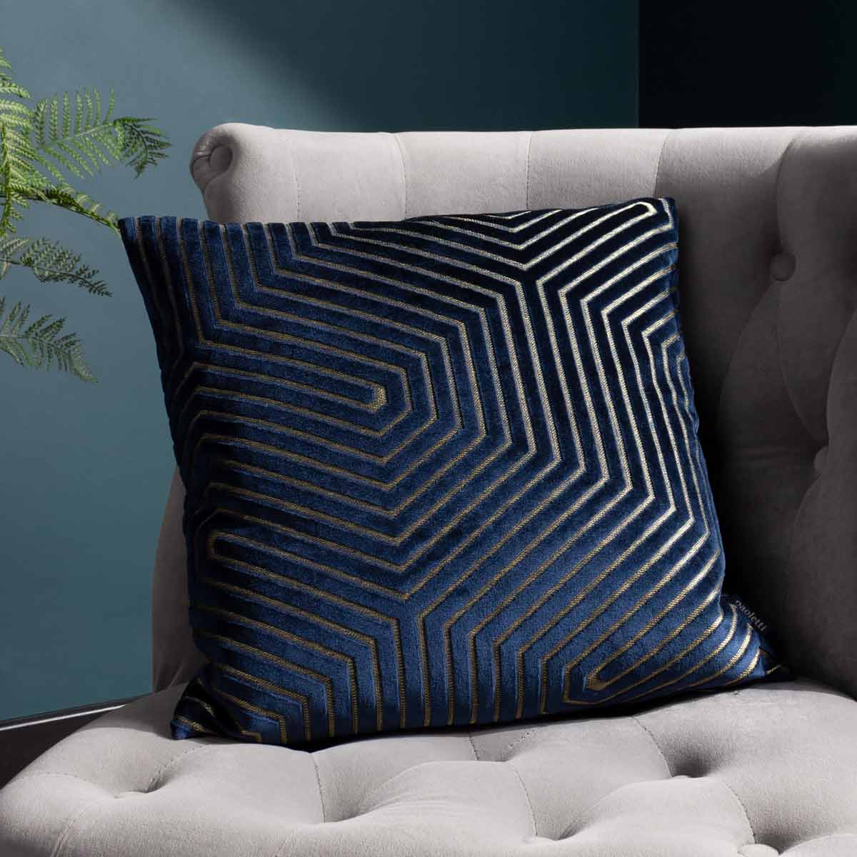 Navy geometric cut velvet cushion featuring a striking pattern of interlocking lines, measuring 45cm x 45cm, set against a soft, upholstered sofa.