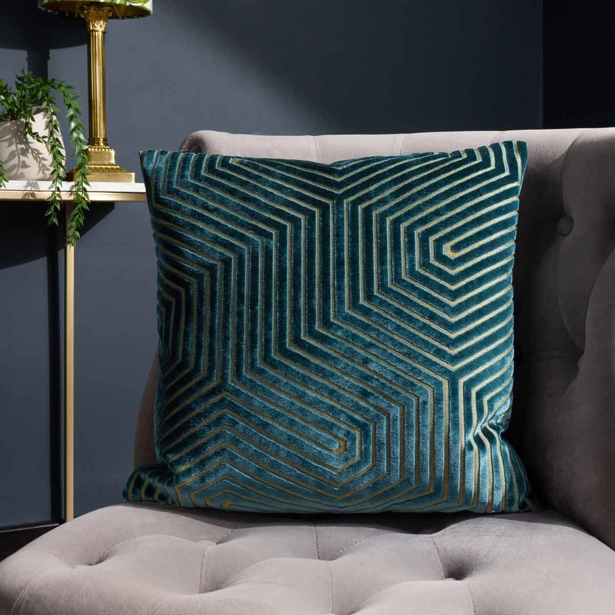 Teal geometric cut velvet cushion featuring a striking linear pattern, measuring 45cm x 45cm, displayed on a plush gray sofa with a stylish lamp in the background.