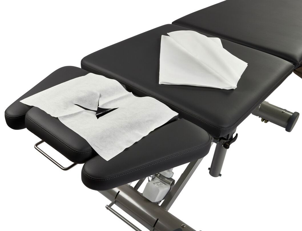 White paper towels are neatly stacked on a black padded treatment table, with one towel placed open, showcasing a cut-out design for easy use.