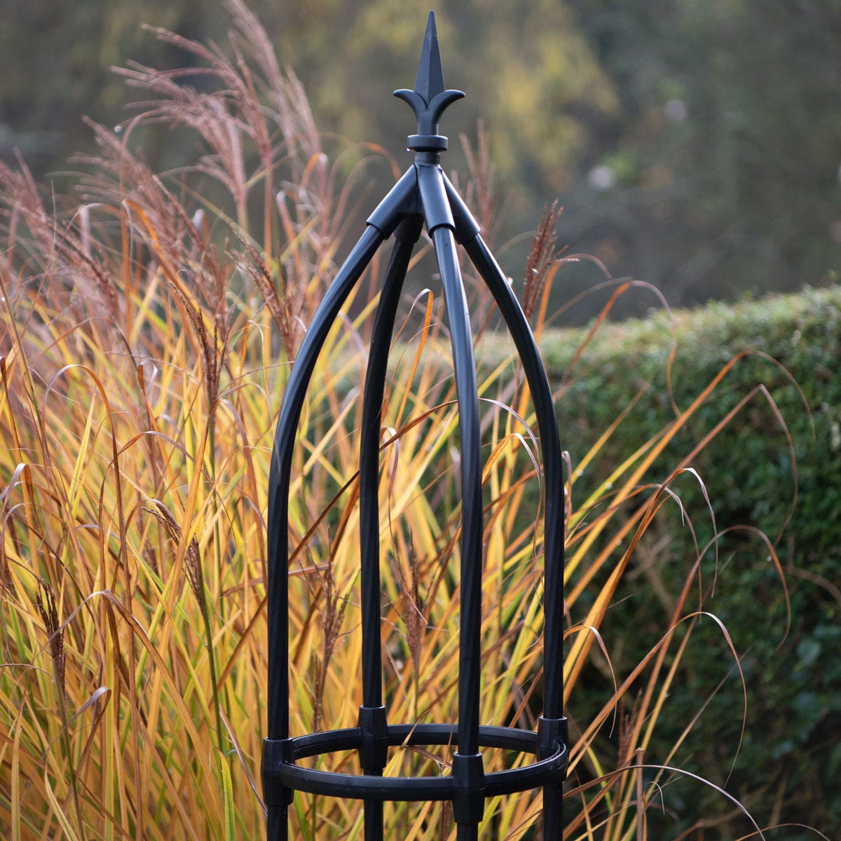 Black metal garden obelisk standing 1.7m tall, featuring a pointed top and a circular base, surrounded by tall golden grasses in a garden setting.