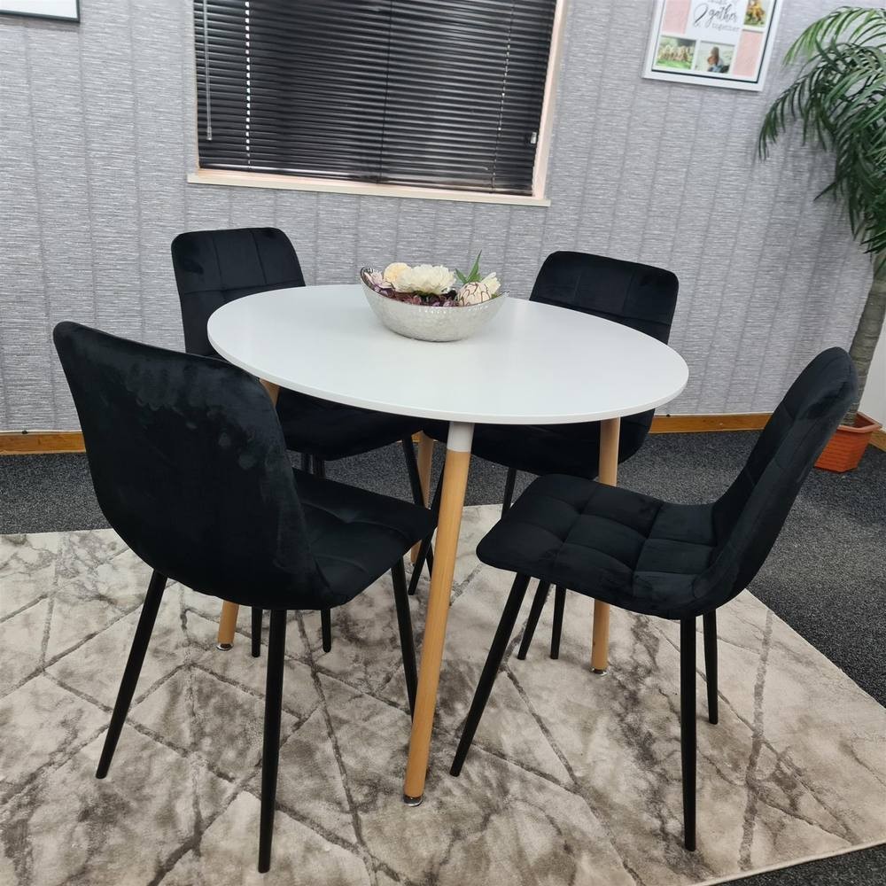 White round wooden dining table surrounded by four black velvet chairs with sleek black legs, set on a patterned rug in a modern room with a plant and decorative bowl centerpiece.