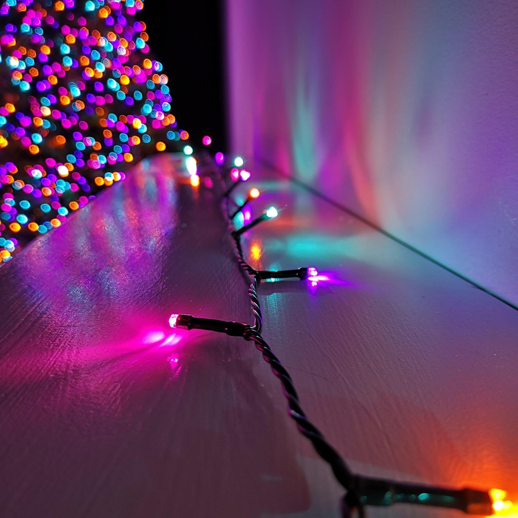 Rainbow-colored LED string lights with a black wire, featuring vibrant purple, blue, green, and orange bulbs, illuminating a surface, with a blurred Christmas tree in the background.
