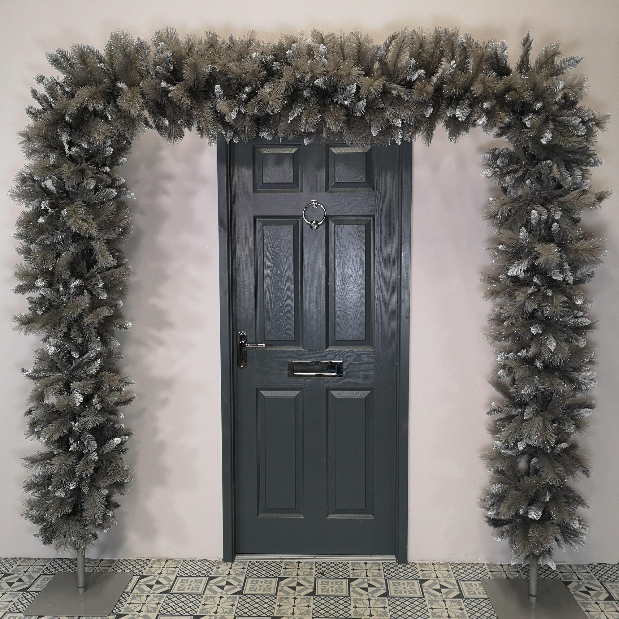 7.5ft silver Christmas tree arch made of PVC branches, featuring 1070 tips with a frosted appearance, placed over a dark door in a patterned floor setting.