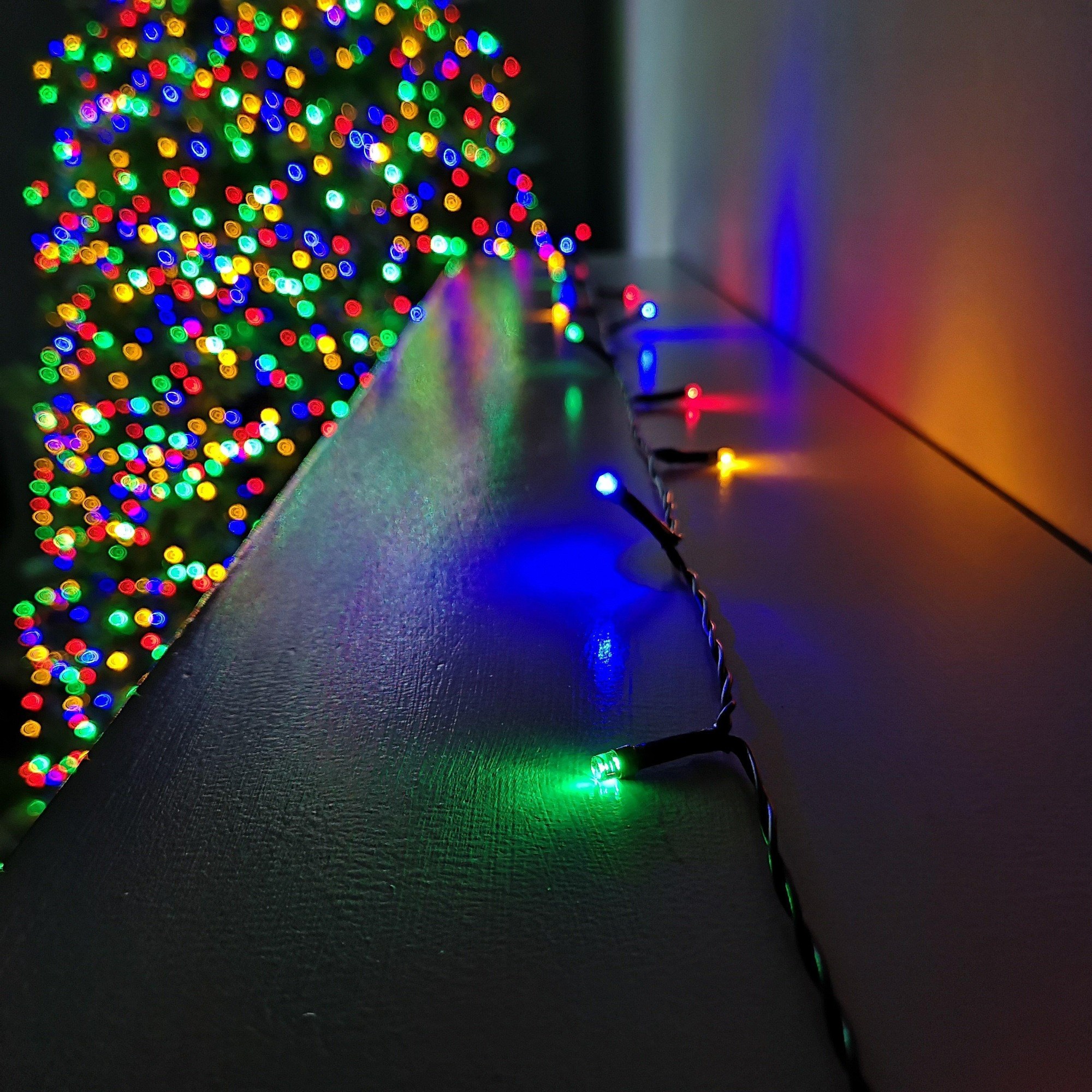 Multicoloured LED string lights with a black wire, featuring blue, green, red, and yellow bulbs, set against a blurred, brightly lit Christmas tree backdrop.