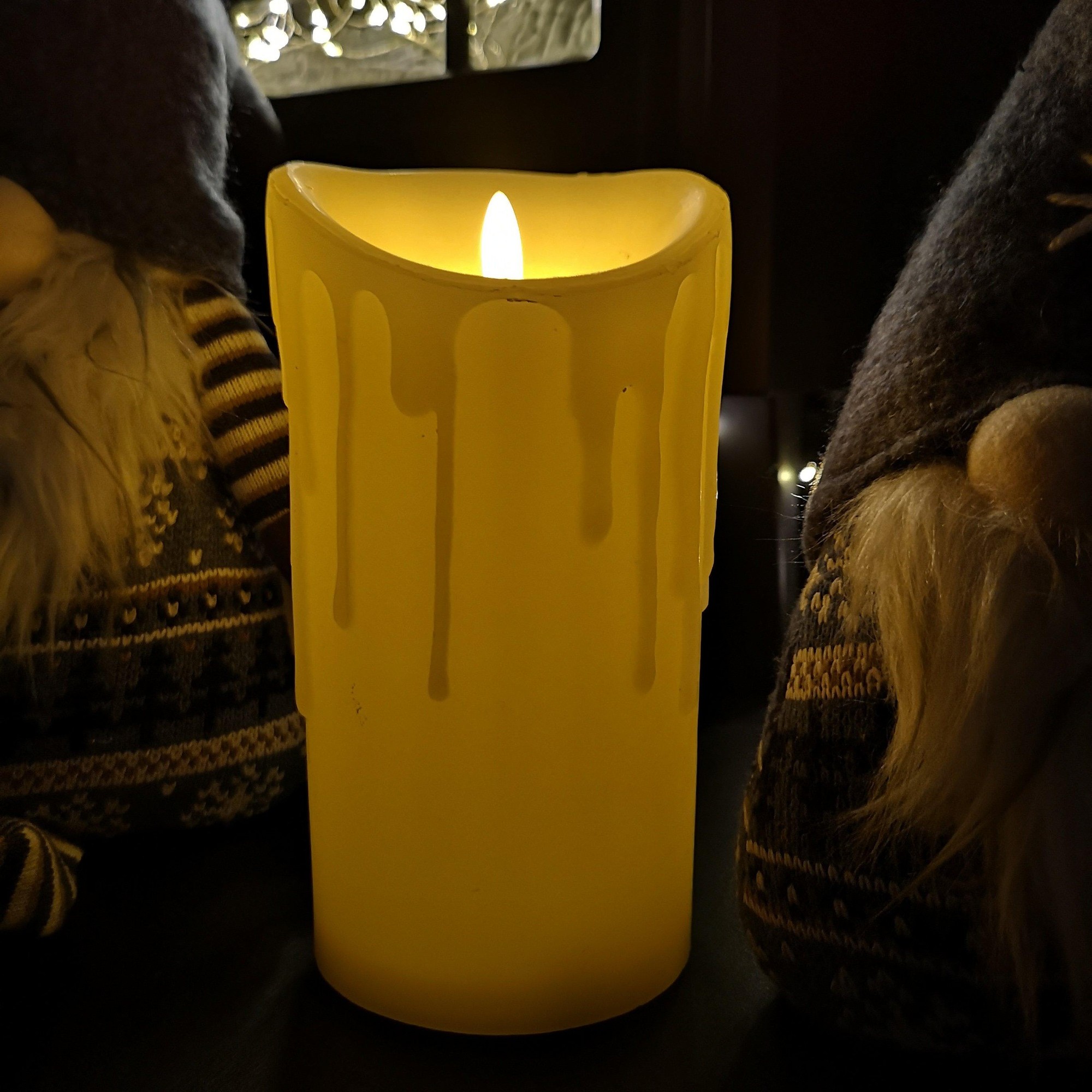 Cream-colored LED candle with a realistic flickering flame and melted effect, measuring 18x9cm, surrounded by decorative figures.