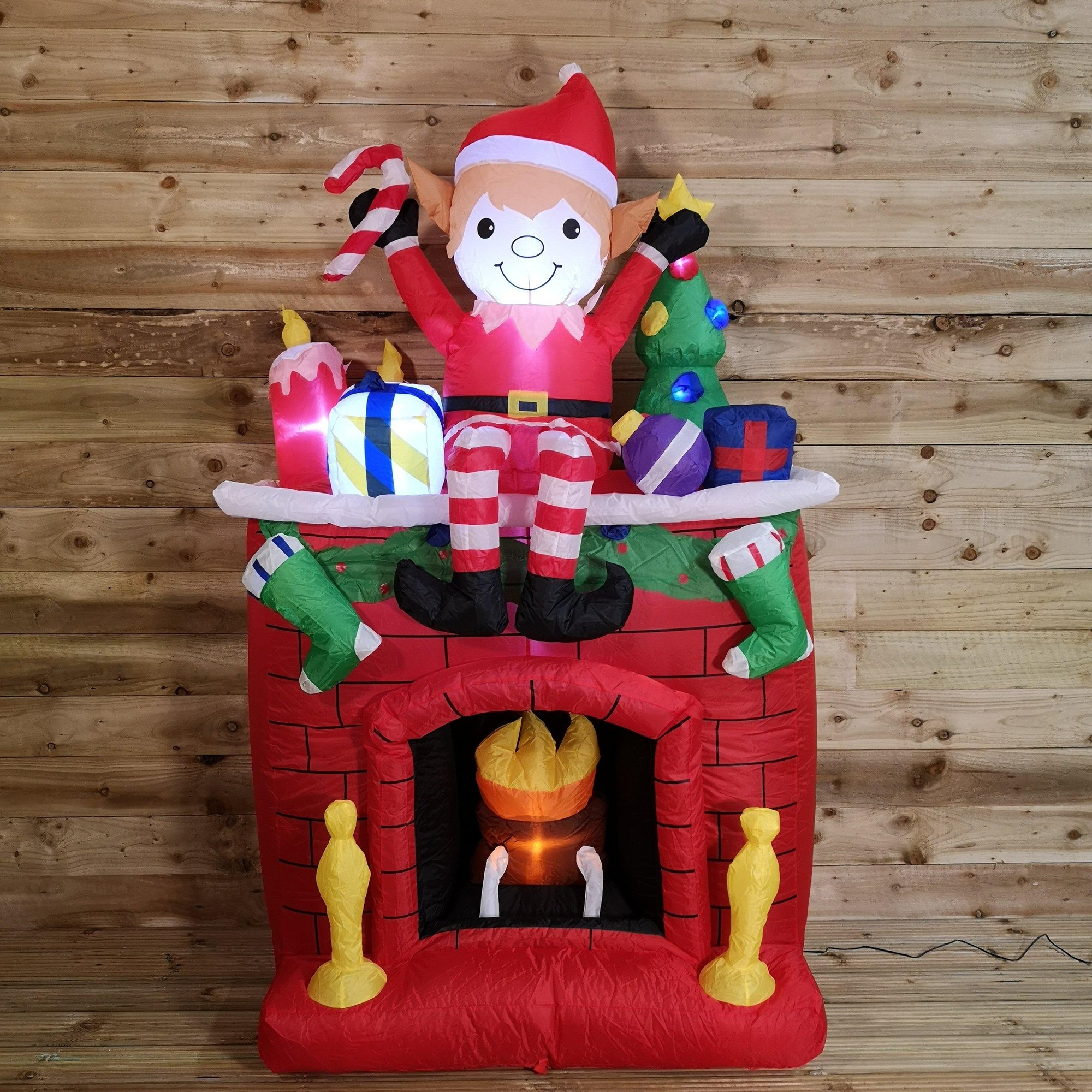 A vibrant inflatable fireplace featuring a cheerful elf in a red outfit, surrounded by colorful presents and stockings, with glowing lights and a warm, flickering flame effect.