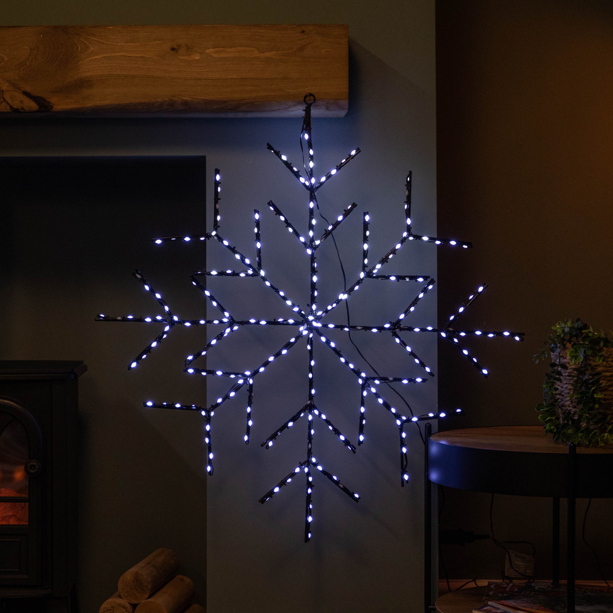 76cm light-up snowflake decoration made of black metal with 250 white LEDs, featuring a geometric design, hanging against a dark wall near a cozy fireplace.