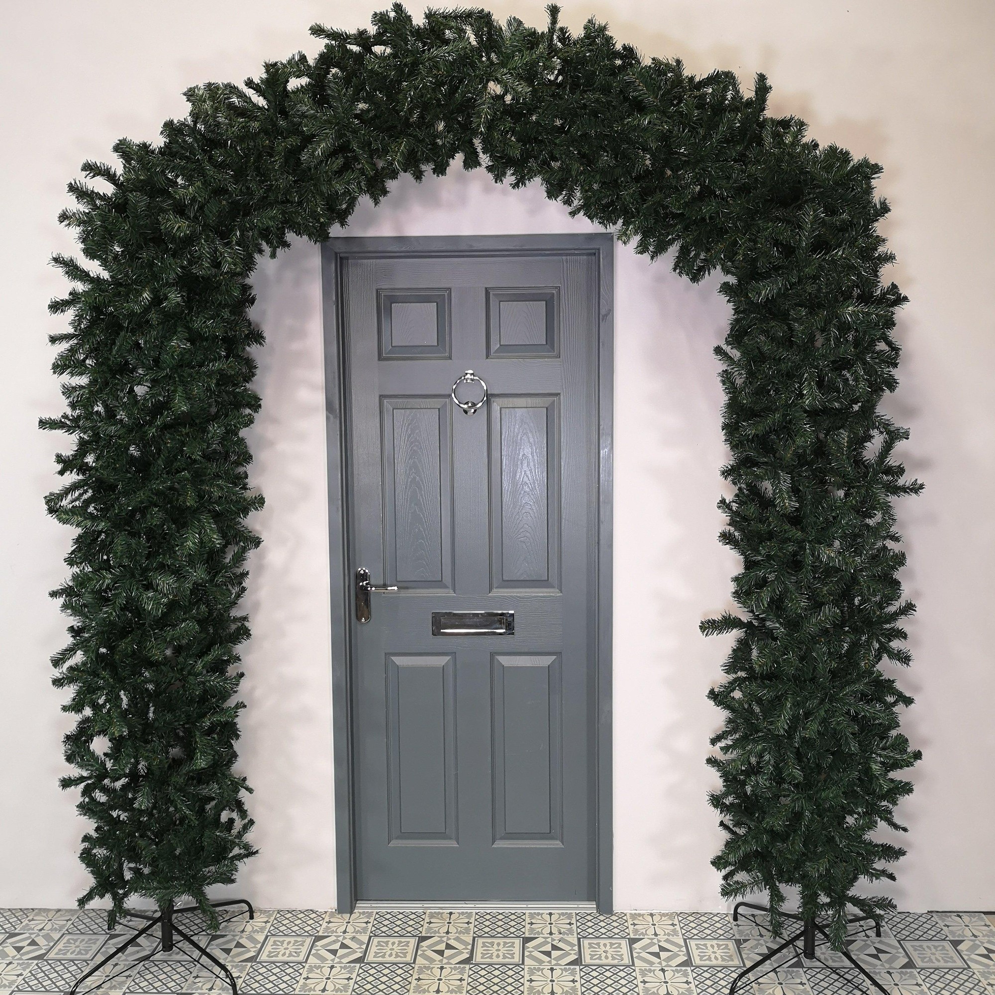 8ft tall green Christmas tree arch made of faux pine branches, featuring a sturdy metal frame and two supporting stands, ideal for indoor or outdoor festive decoration.