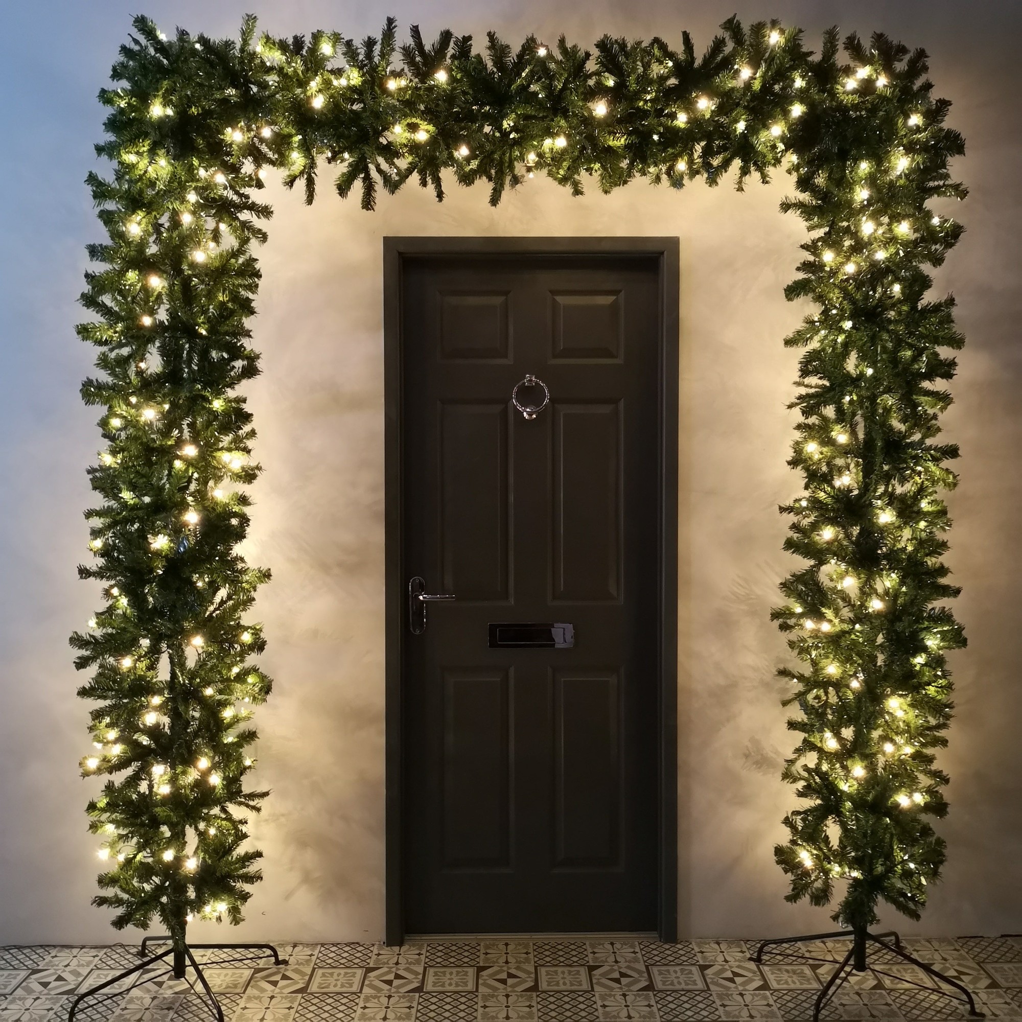 8ft tall green prelit Christmas tree arch with lush foliage and warm white lights, framing a dark door against a light wall and patterned floor.