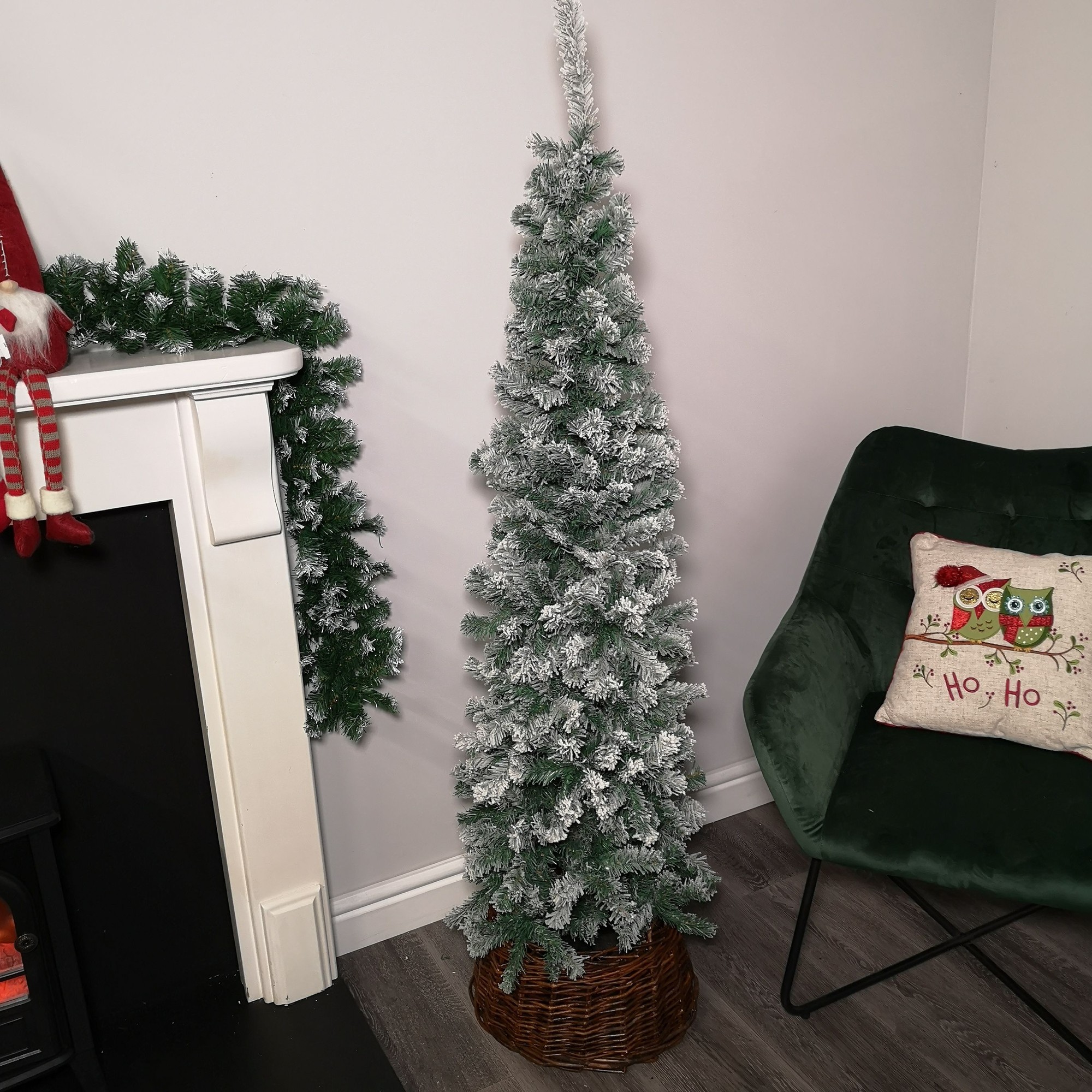 Slim 5.5ft green Christmas tree with snow-flocked branches, set in a woven basket, positioned next to a fireplace and a green chair, decorated with a festive pillow.