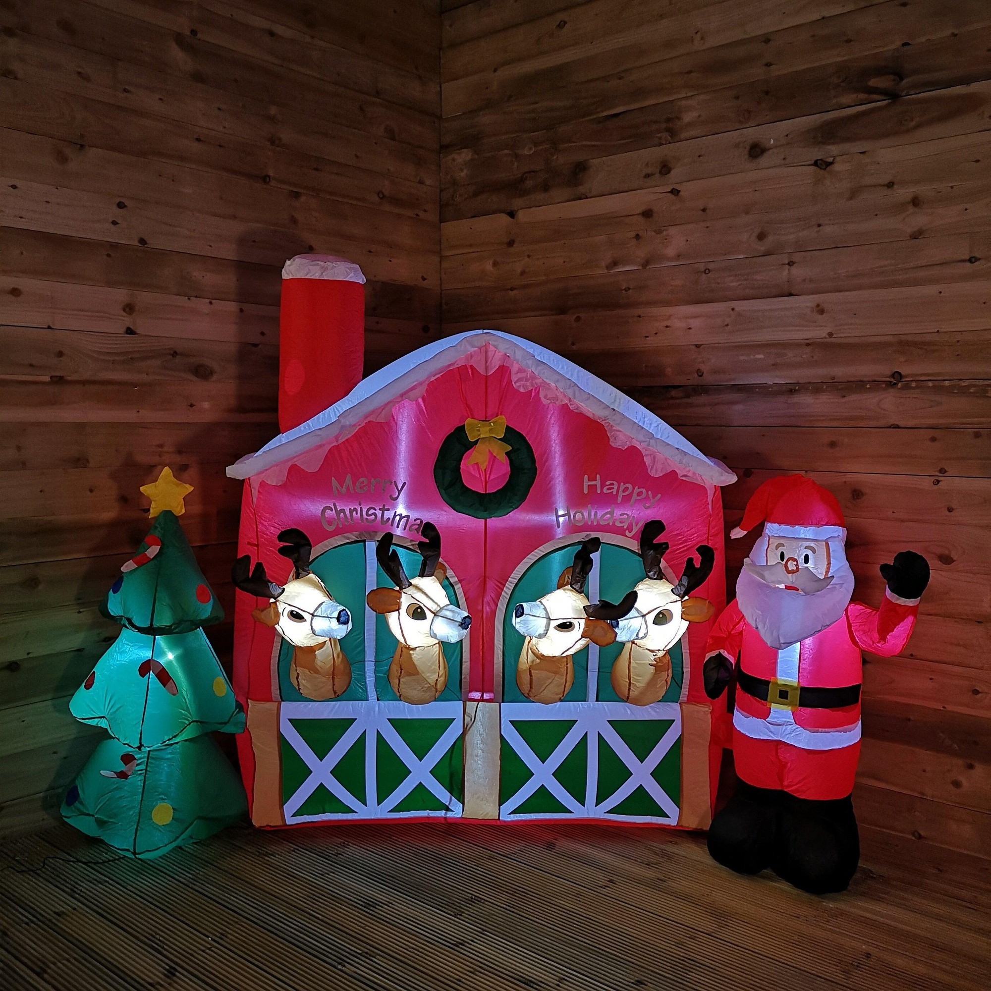 A colorful 2.7M inflatable Christmas stable featuring Santa, reindeer, and a Christmas tree, with bright lights and festive decorations on a wooden background.