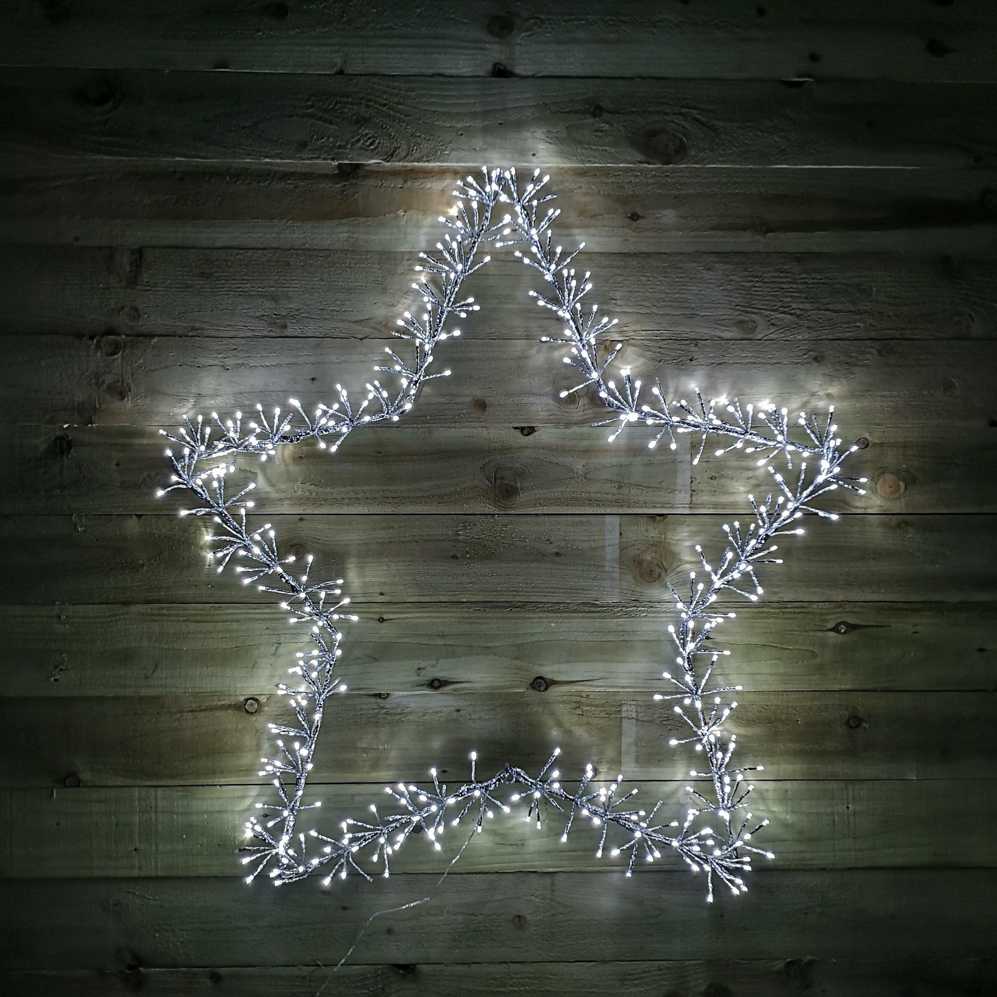 A silver star shape made of flexible branches adorned with 480 white LED lights, designed for both indoor and outdoor festive decoration against a wooden background.