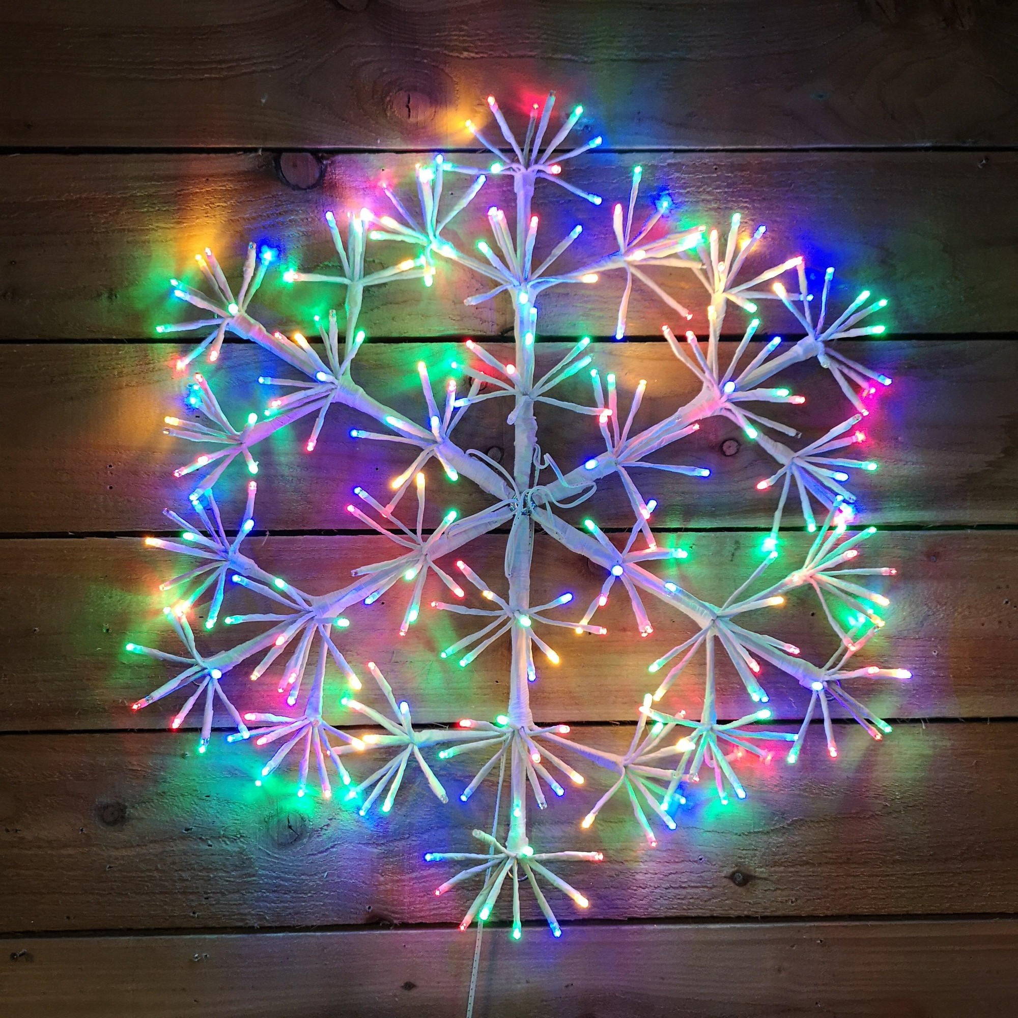 60cm white starburst snowflake made of plastic with multi-coloured twinkling LED lights, featuring vibrant red, blue, green, and yellow hues against a wooden background.