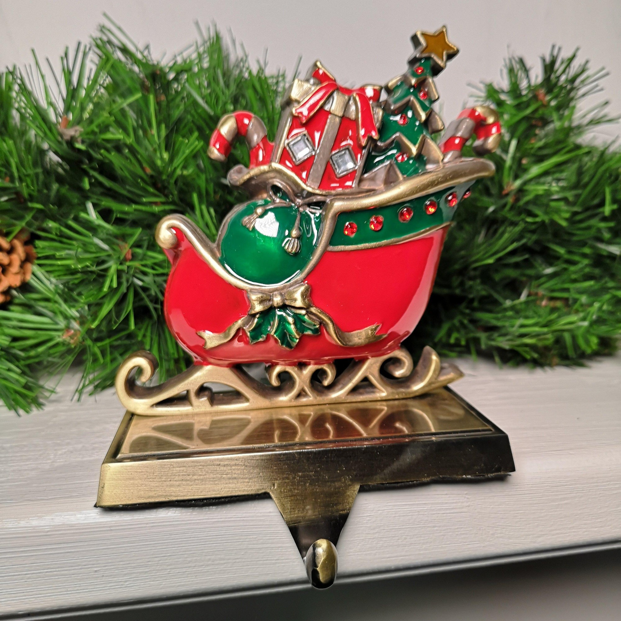 Red and green sleigh stocking hanger with candy canes, presents, and a Christmas tree, featuring gold accents and a sturdy base for hanging.