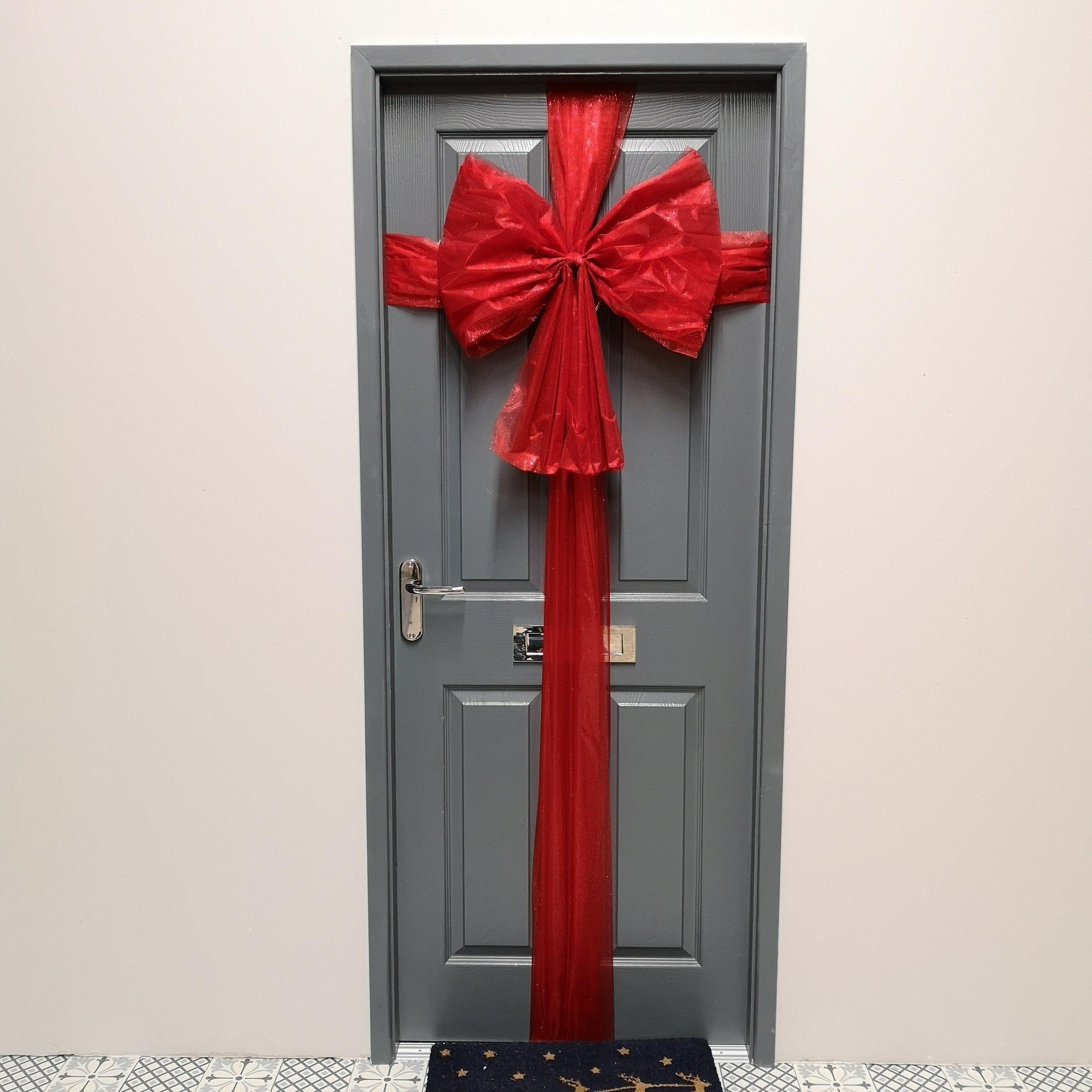 Large red bow with a matching ribbon wrap adorns a grey door, creating a festive Christmas decoration. The bow features sheer fabric for a shimmering effect.
