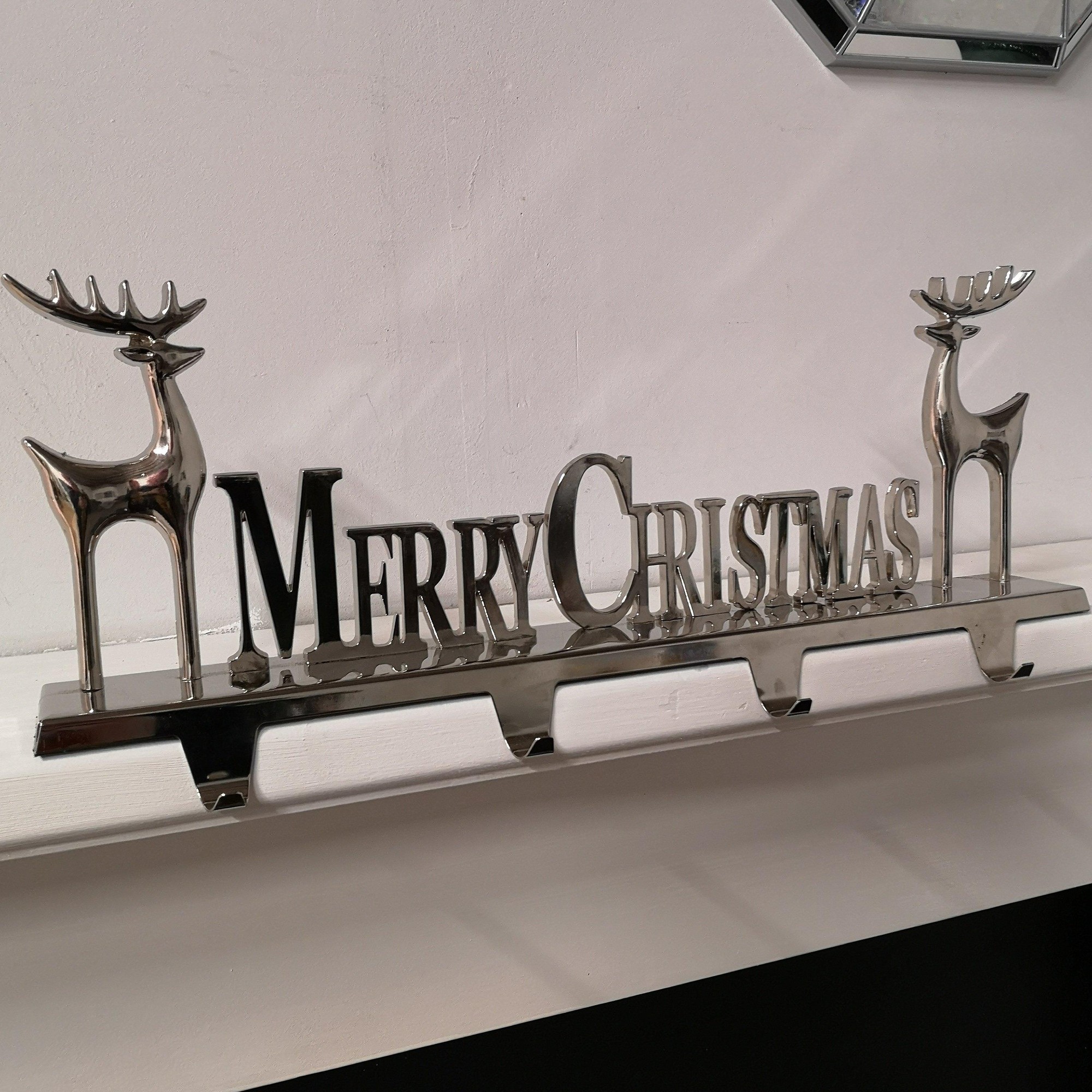 Silver decorative stocking hanger featuring "MERRY CHRISTMAS" text and two reindeer designs, designed to hold four stockings, with a polished metallic finish.